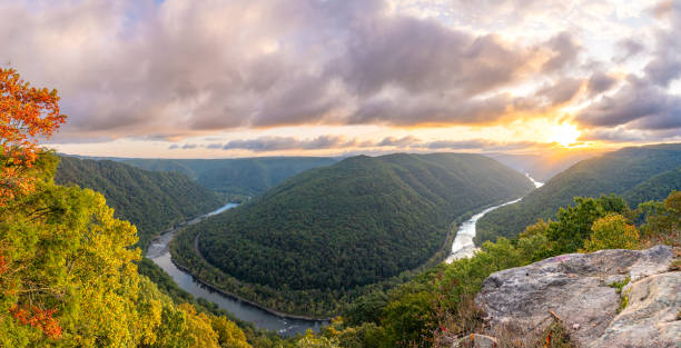 Panoramic view of a winding river cutting through lush, green forested hills at sunrise, with dramatic clouds and sunlight in the sky—an ideal scene for day trips from Berkeley Springs, seen from a rocky overlook.