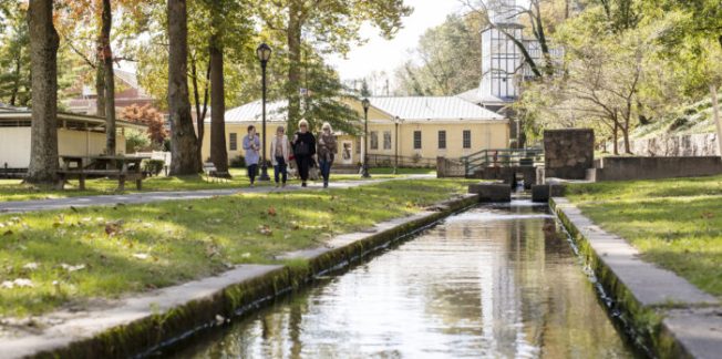 A narrow stream runs through a grassy park with tall trees. Four people walk along a path beside the stream, and historic-looking yellow buildings stand in the background under a clear sky.